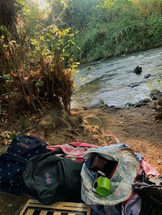 there is a backpack and other items on the ground next to a river with trees in the background