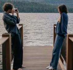 a man and woman standing on a dock looking at the water with mountains in the background