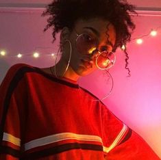 a woman with curly hair and round glasses standing in front of a pink wall wearing hoop earrings