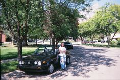 a woman standing next to a parked car on the side of a road near trees