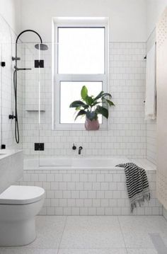 a white tiled bathroom with a toilet, tub and window in the corner that has a potted plant next to it