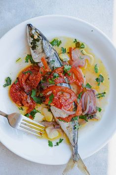 a white plate topped with fish covered in tomatoes and onions next to a fork on top of a table