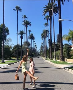 two women are crossing the street in front of palm trees and one is holding her hand up