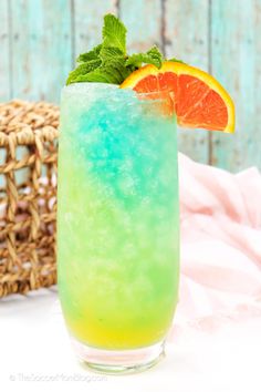 a tall glass filled with blue and green liquid next to a basket full of orange slices