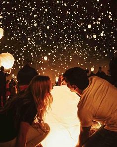 two people looking at paper lanterns in the sky with lights all around them and onlookers watching