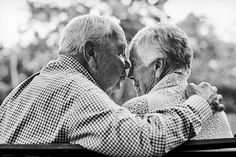 two elderly people sitting on a bench in the park, one holding his arm around the other's neck