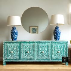 a blue and white sideboard with two lamps on it next to a round mirror