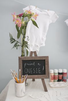 an easel with paint, pencils and flowers in it next to a sign that says paint a onesie