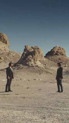 two men in suits are standing in the middle of an arid area with rocks on either side