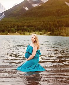 a pregnant woman in a blue dress sitting on the edge of a body of water