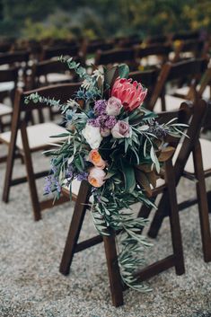 an arrangement of flowers and greenery is placed on the back of wooden folding chairs