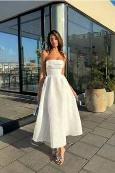 a woman in a white dress is standing on the sidewalk near a building and looking at the camera