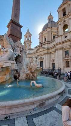 a fountain in front of a large building with statues on it and people walking around