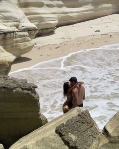 a man and woman kissing on the beach next to some rocks with waves coming in