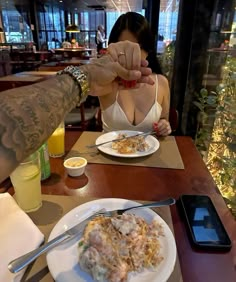 a woman sitting at a table with plates of food in front of her and a cell phone