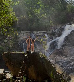 two women in bikinis standing on top of a rock next to a water fall