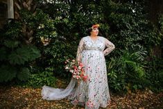 a woman standing in front of some trees and bushes wearing a dress with flowers on it