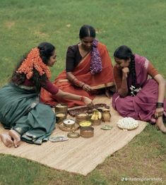 three women are sitting on a blanket in the grass and having a picnic with food