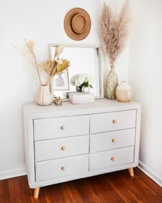 a white dresser topped with lots of drawers next to a mirror and vase filled with flowers