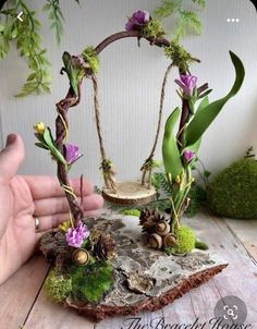 a hand holding a miniature swing with flowers and plants on it, sitting on top of a piece of wood