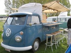 an old vw camper van is parked in the grass with tables and chairs