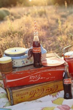 an old coca - cola cooler and other items are sitting on a blanket in the middle of a field