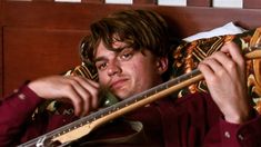 a young man laying in bed with a guitar on his shoulder and looking at the camera