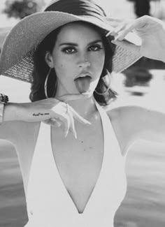 a woman in a white dress and hat sticking her tongue out while standing next to a body of water