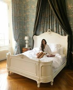 a woman sitting on top of a white bed in a bedroom next to a window