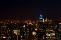 new york city at night with the empire building lit up in blue and white, as seen from top of the rock