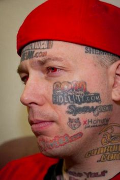 a man with tattoos on his face and neck, looking to the side while wearing a red baseball cap