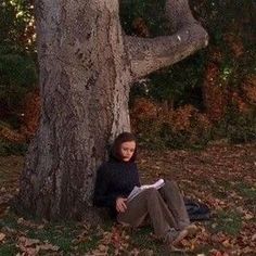 a woman sitting on the ground next to a tree with an elephant standing behind her