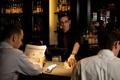 three men are sitting at a bar and one is reading the paper while the other man looks on