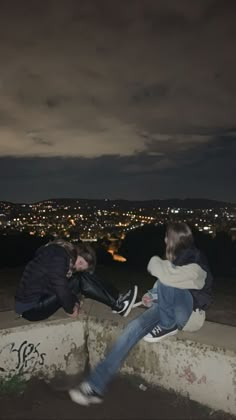 two people sitting on top of a cement wall with the city lights in the background