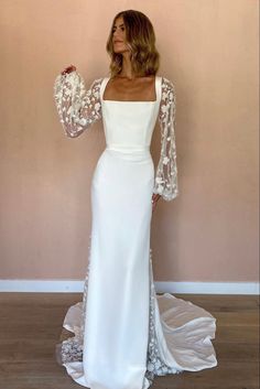 a woman standing in front of a wall wearing a long white dress with sheer sleeves