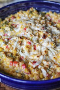 a blue bowl filled with corn and cheese on top of a wooden table next to bread