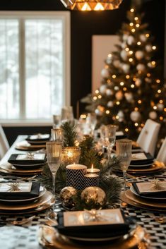 a dining room table set with black and white plates, silverware, candles and a christmas tree in the background