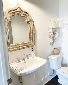 An antiqued cathedral mirror with a thick wood frame is hung over a vintage pedestal sink beside a matching distressed floating wood shelf. Decorate this cool neutral space with a woven toiletry basket modern black soap dispenser, white foliage, and a farmhouse pinstriped hand towel...   Image: white.cottage.life