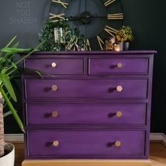 a purple dresser with gold knobs sits in front of a clock and houseplants