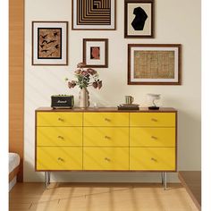 a yellow dresser sitting in front of a wall with pictures on it and vase filled with flowers