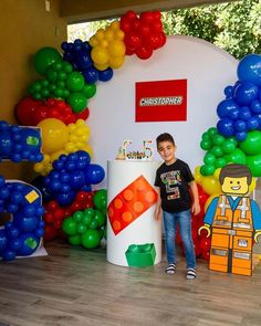 a young boy standing in front of a giant lego cake and balloon arch at a birthday party