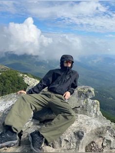 a man sitting on top of a large rock next to a lush green forest under a blue cloudy sky