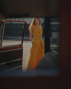 a woman in a yellow dress standing next to a red car and looking at the camera