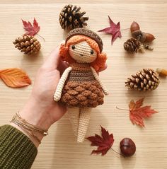 a hand is holding a knitted doll surrounded by autumn leaves and pinecones