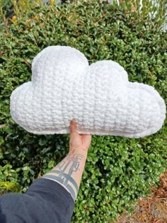 a hand is holding up a crocheted cloud pillow in front of some bushes