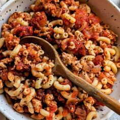 a white bowl filled with pasta and meat next to a wooden spoon on top of a table