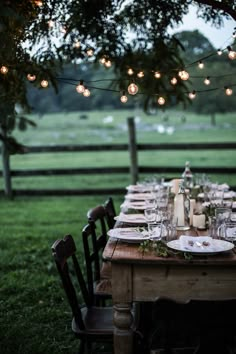 an outdoor dinner table set up with plates and wine glasses on it, surrounded by string lights