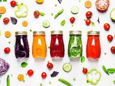 several jars filled with different colored liquid surrounded by various vegetables and fruits on a white surface