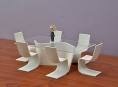 a glass table with four chairs and a vase on it, sitting in front of a purple wall