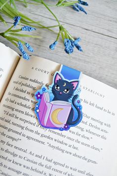 an open book with a black cat on it next to blue flowers and green leaves